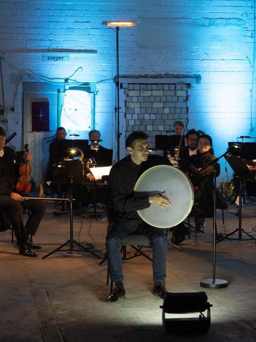 Auf einer Bühne in einem blau ausgeleuchtetem Industrieraum sitzt ein auf Abstand bedachtes Orchester, im Vordergrund ein Mann mit großer runder Trommel.