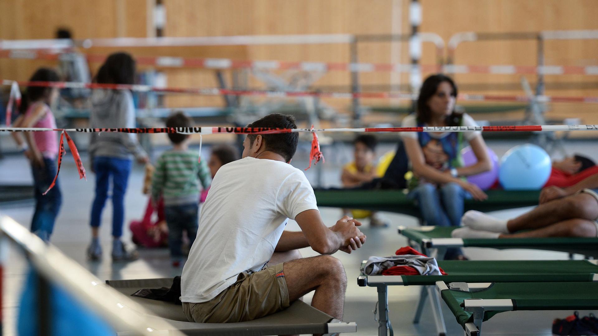 Flüchtlinge sitzen am 04.08.2015 auf Feldbetten in einer Turnhalle auf dem Gelände der Bundespolizei in Rosenheim. Flüchtlinge sitzen am 04.08.2015 auf Feldbetten in einer Turnhalle auf dem Gelände der Bundespolizei in Rosenheim.