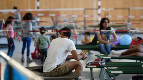Flüchtlinge sitzen am 04.08.2015 auf Feldbetten in einer Turnhalle auf dem Gelände der Bundespolizei in Rosenheim.