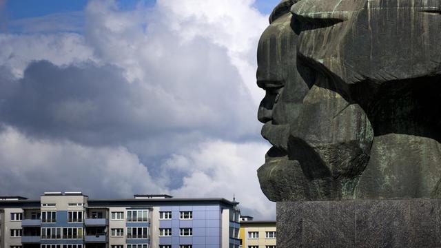 Karl-Marx-Denkmal in Chemnitz, dahinter ein Hausdächer und ein bewölkter Himmel.