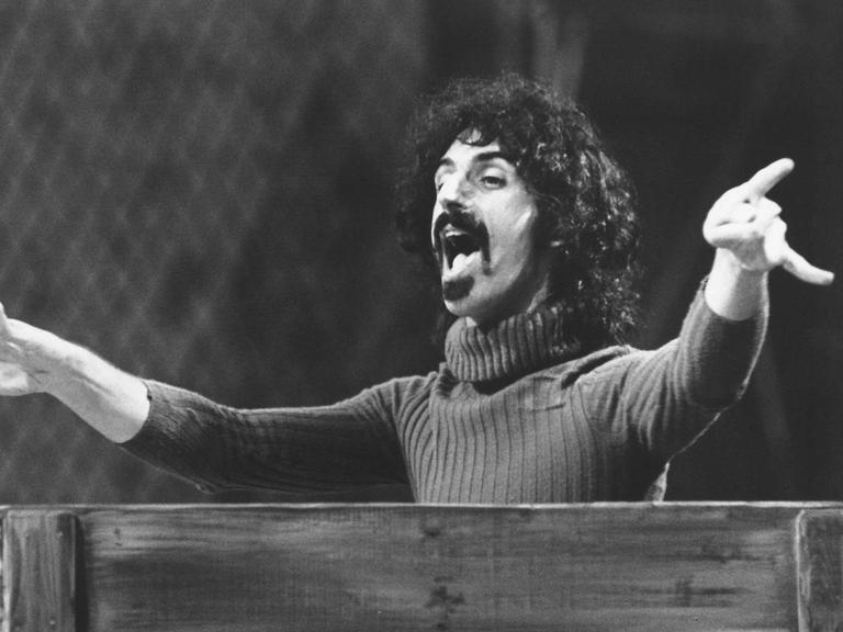 Frank Zappa steht hinter einem Pult in Dirigentenpose.