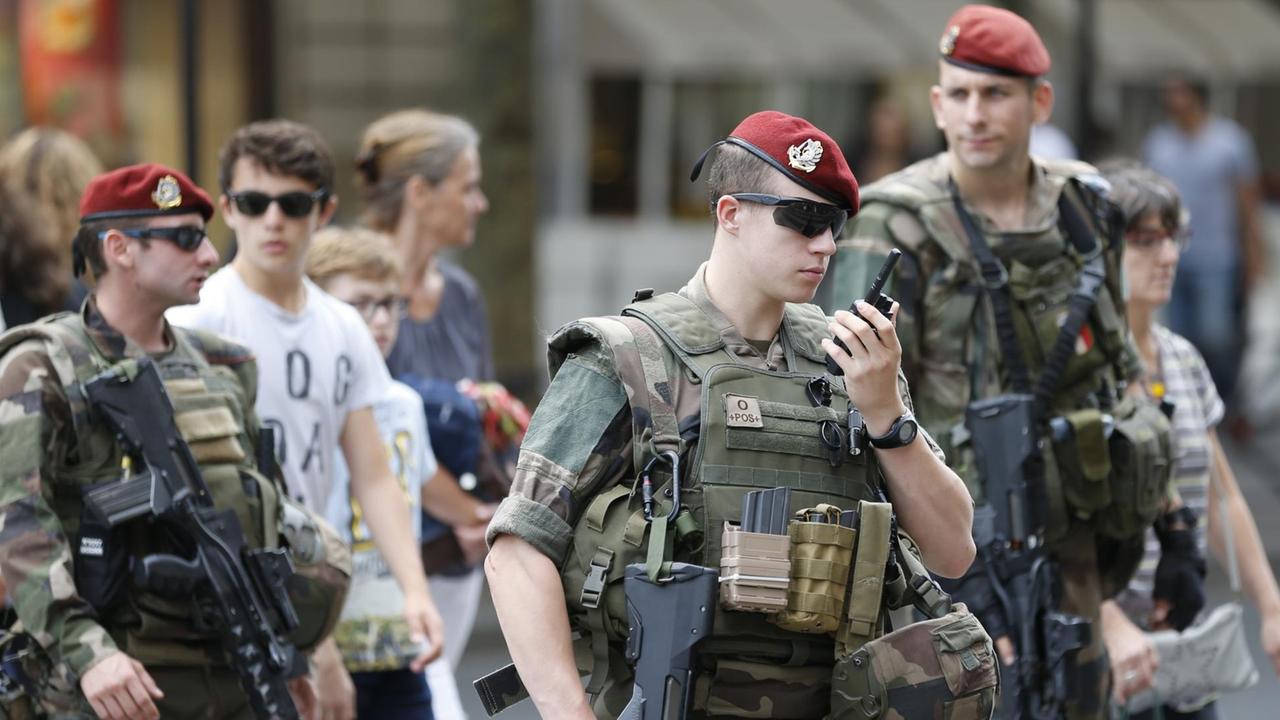 Drei Soldaten mit roten Barretten und Maschinengewehren; zwei von ihnen tragen Sonnenbrillen. Einer spricht in ein Funkgerät.
