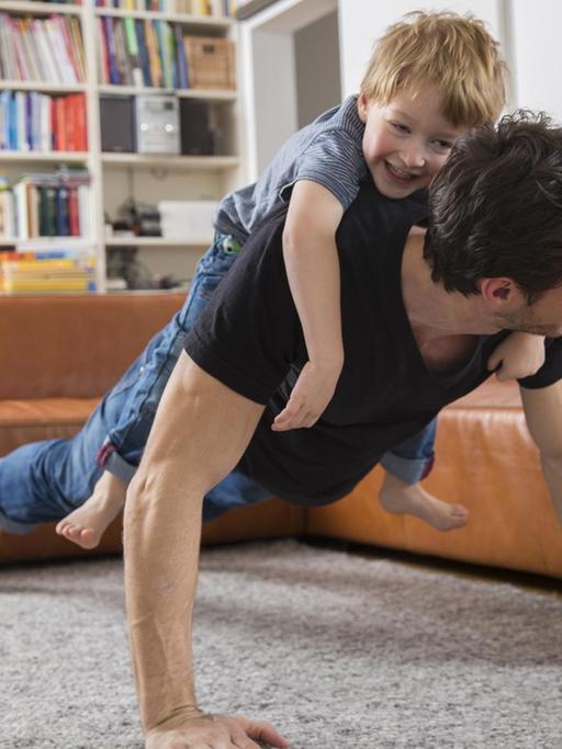 Ein Vater macht Liegestütze im Wohnzimmer mit seinem Sohn auf dem Rücken.