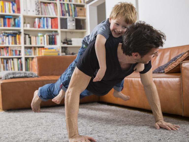 Ein Vater macht Liegestütze im Wohnzimmer mit seinem Sohn auf dem Rücken.