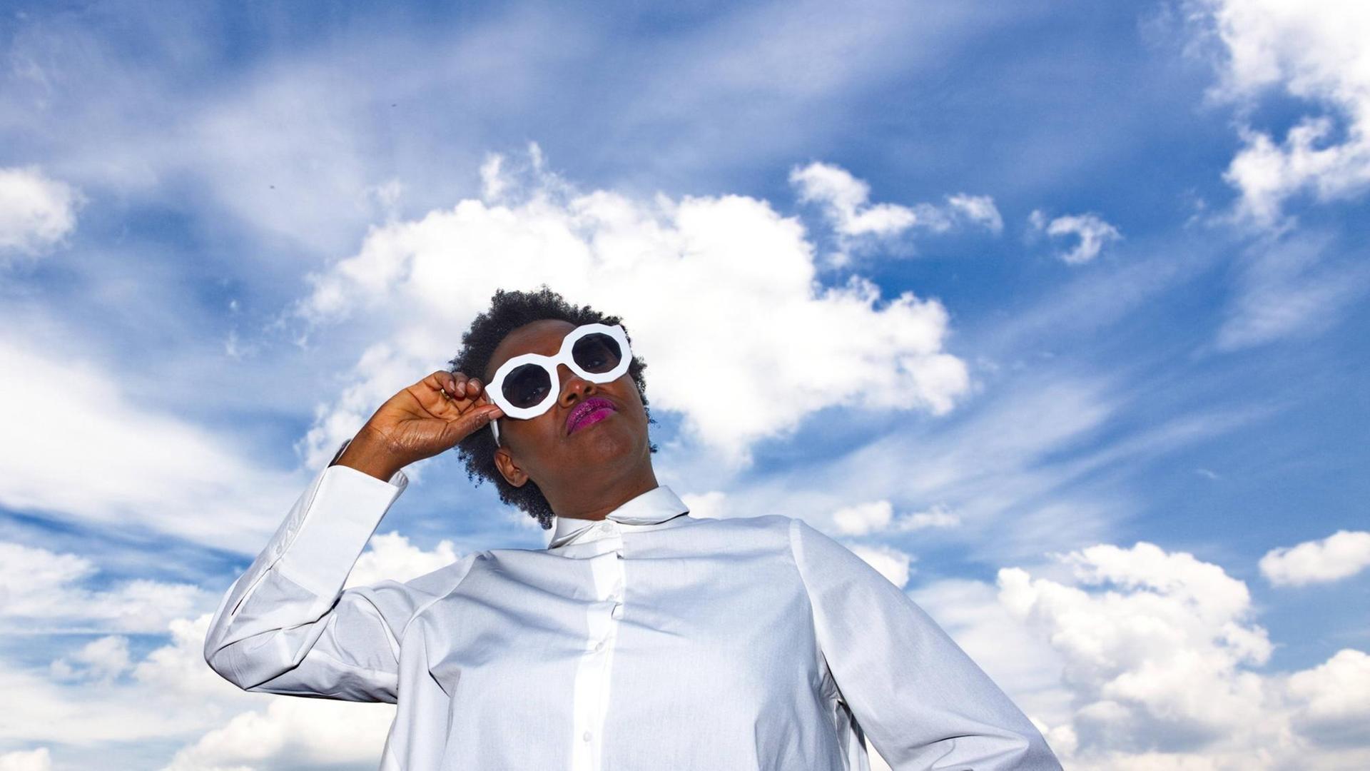 Onejiru steht auf einem Dach, trägt ein weißes Kleid und Sonnebrille — hinter ihr der blaue Himmel mit weißen Wolken Onejiru steht auf einem Dach, trägt ein weißes Kleid und Sonnebrille — hinter ihr der blaue Himmel mit weißen Wolken