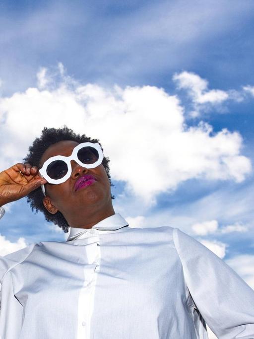 Onejiru steht auf einem Dach, trägt ein weißes Kleid und Sonnebrille — hinter ihr der blaue Himmel mit weißen Wolken