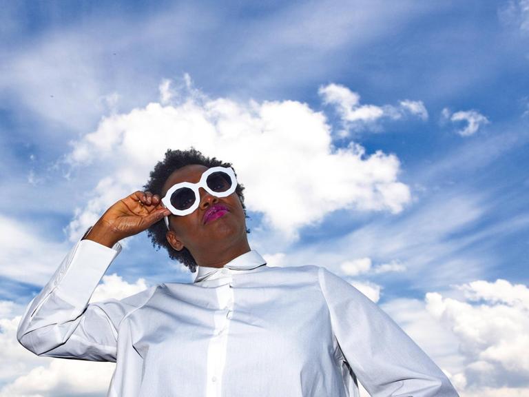 Onejiru steht auf einem Dach, trägt ein weißes Kleid und Sonnebrille — hinter ihr der blaue Himmel mit weißen Wolken