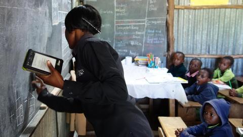 An "Bridge"-Schulen unterrichten die Lehrerinnen und Lehrer unterstützt durch Tablet-Computer. Hier eine Lehrerin in Kenia, die an der Tafel mit Kreide steht und in der anderen Hand ein Tablet hält.