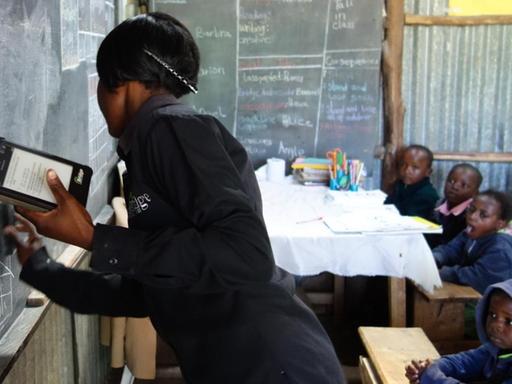 An "Bridge"-Schulen unterrichten die Lehrerinnen und Lehrer unterstützt durch Tablet-Computer. Hier eine Lehrerin in Kenia, die an der Tafel mit Kreide steht und in der anderen Hand ein Tablet hält.