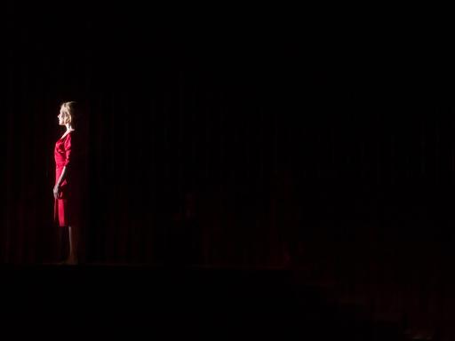 Eine Frau in einem roten Kleid auf der Bühne vor einem roten Vorhang.