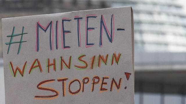 Ein hochgehaltenes Pappschild mit der Aufschrift "#Mietenwahnsinn stoppen". Im Hintergrund ist die Kuppel des Reichstags zu sehen.