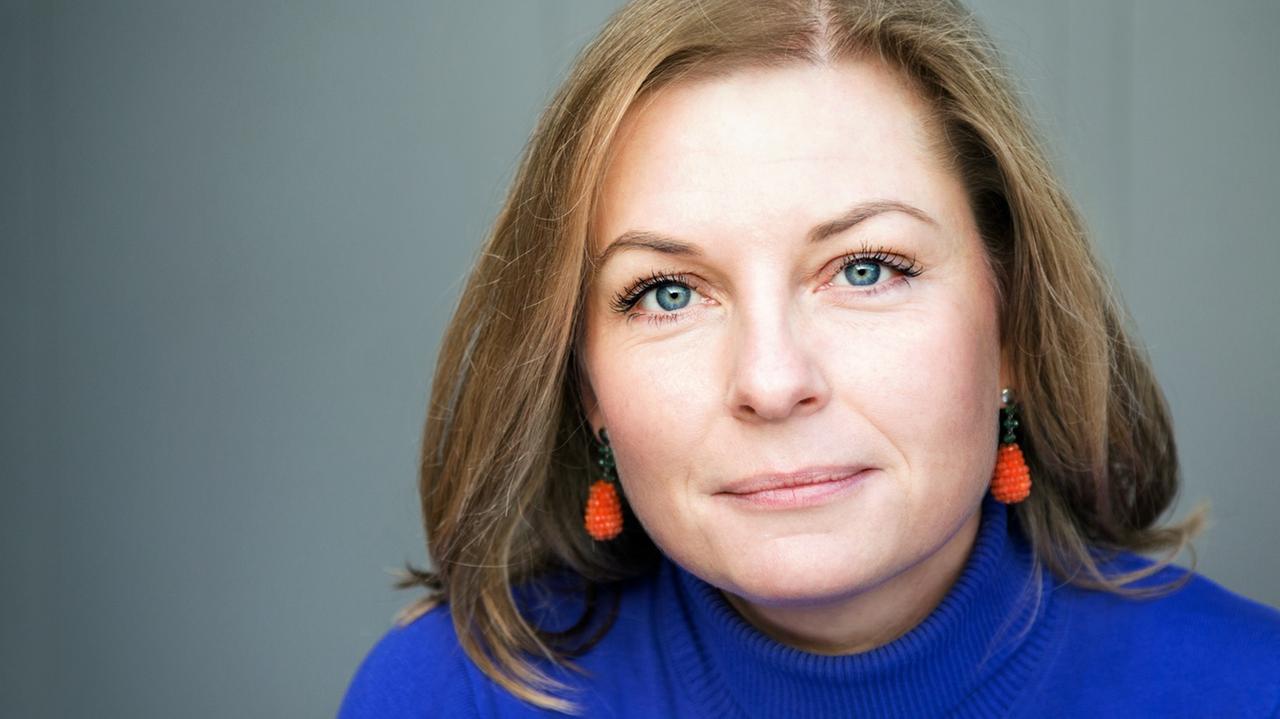 Sandra Anusiewicz-Baer trägt einen blauen Pullover und orange Ohrringe. Sandra Anusiewicz-Baer trägt einen blauen Pullover und orange Ohrringe.