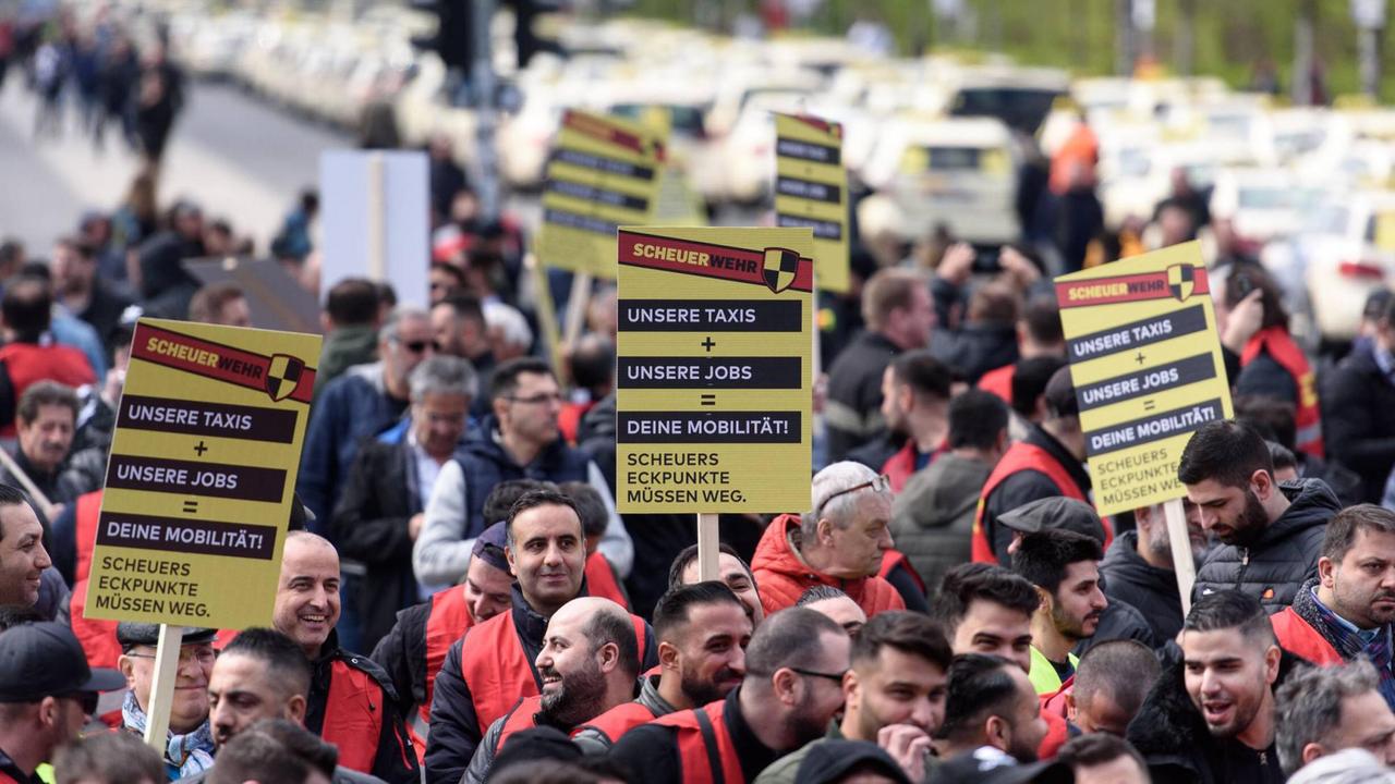 Im Bild sind streikende Taxifahrer vor der Bühne mit Schildern zu sehen. Die Schilder tragen die Aufschrift "Unsere Taxis und unsere Jobs sind deine Mobilität! Scheuers Eckpunkte müssen weg." Im Bild sind streikende Taxifahrer vor der Bühne mit Schildern zu sehen. Die Schilder tragen die Aufschrift "Unsere Taxis und unsere Jobs sind deine Mobilität! Scheuers Eckpunkte müssen weg."