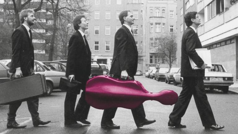 Vier Musiker mit verschiedenen Instrumenten laufen hintereinander über die Straße in Richtung Probenort