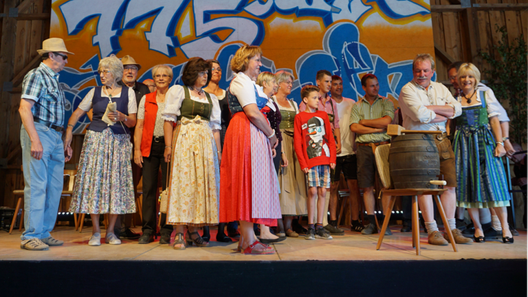 Auf der Bühne stehen Frauen und Männer in traditioneller Kleidung, auf einem Stuhl ein Bierfass. Auf der Bühne stehen Frauen und Männer in traditioneller Kleidung, auf einem Stuhl ein Bierfass.