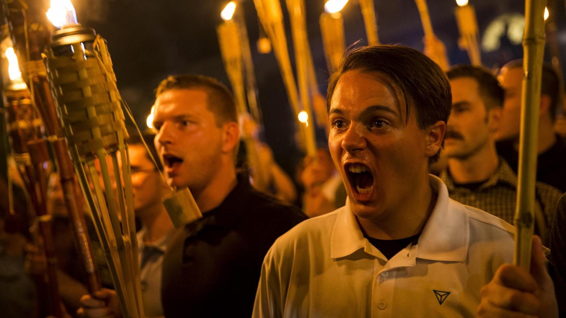 Demonstration von Neonazis und Mitgliedern der Alt-Right-Bewegung im August 2017 in Charlottesville, VA. Junge Männer halten im Dunkeln brennende Fackeln in der Hand und schreien etwas. Demonstration von Neonazis und Mitgliedern der Alt-Right-Bewegung im August 2017 in Charlottesville, VA. Junge Männer halten im Dunkeln brennende Fackeln in der Hand und schreien etwas.