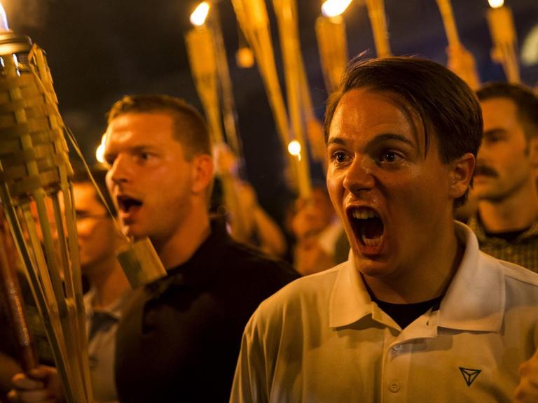 Demonstration von Neonazis und Mitgliedern der Alt-Right-Bewegung im August 2017 in Charlottesville, VA. Junge Männer halten im Dunkeln brennende Fackeln in der Hand und schreien etwas.