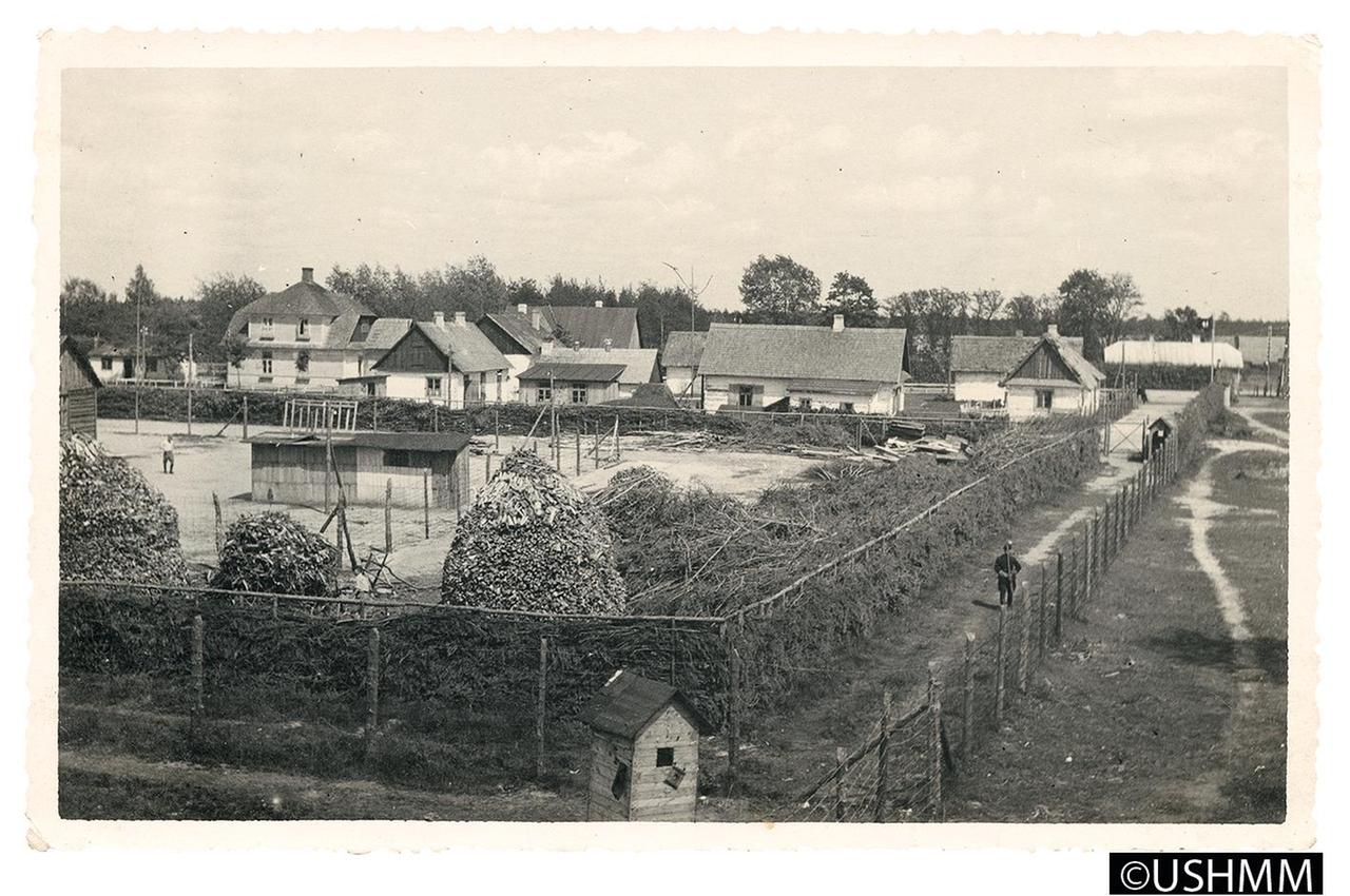 Von einem Wachturm aufgenommene Übersicht über Lager I und das Vorlager im Hintergrund, Frühsommer 1943. Neben der Baracke am linken Bildrand und im Vordergrund zwischen den Brennholz-Stapeln ist jeweils ein jüdischer Zwangsarbeiter zu erkennen. Rechts im Durchgang zwischen den Lagerzäunen patrouillieren zwei Trawniki, in der Verlängerung der Umzäunung ist das helle Dach des Bahnhofsgebäudes sichtbar. Von einem Wachturm aufgenommene Übersicht über Lager I und das Vorlager im Hintergrund, Frühsommer 1943. Neben der Baracke am linken Bildrand und im Vordergrund zwischen den Brennholz-Stapeln ist jeweils ein jüdischer Zwangsarbeiter zu erkennen. Rechts im Durchgang zwischen den Lagerzäunen patrouillieren zwei Trawniki, in der Verlängerung der Umzäunung ist das helle Dach des Bahnhofsgebäudes sichtbar.