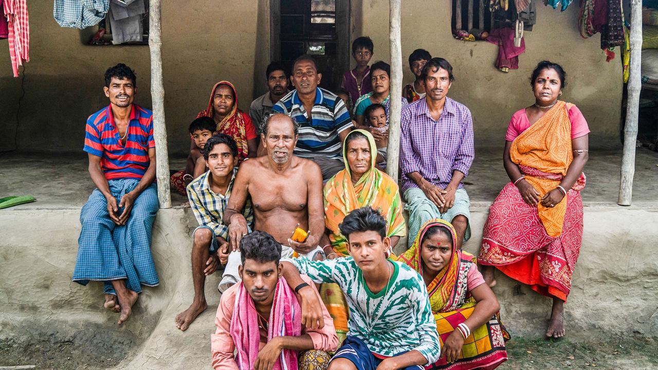 Vor einem Haus sitzt eine Gruppe von Menschen in bunten indischen Gewändern. Im Hintergrund ist Wäsche aufgehängt.