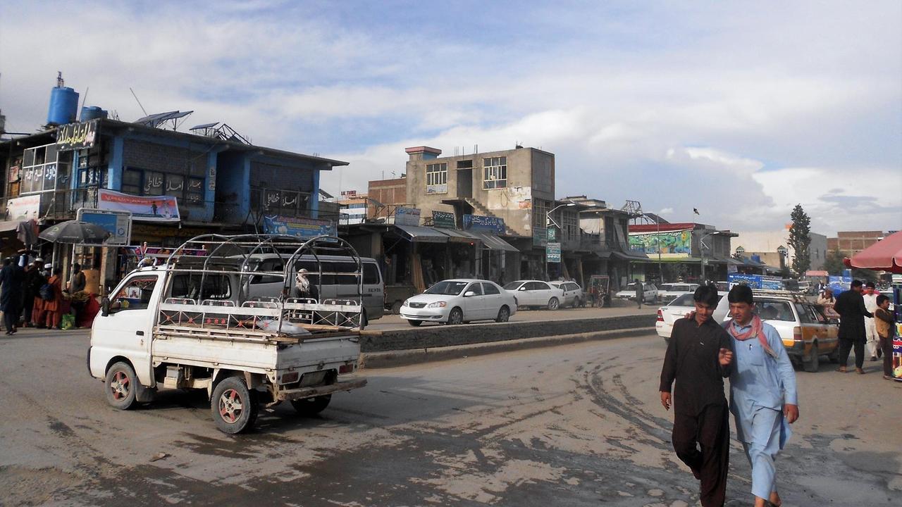 Ein kleiner Lastwagen rollt über die Straße in Khost. Zwei Männer laufen in langen Gewändern daneben.