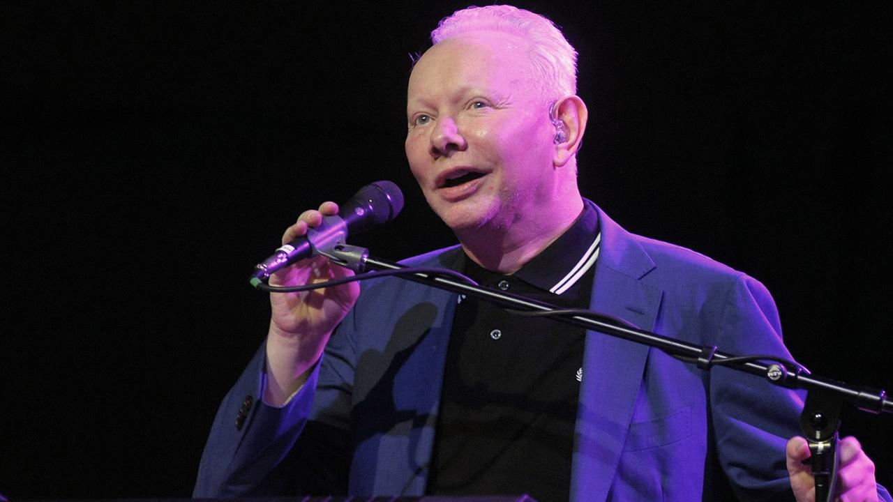 Joe Jackson steht lächelnd am Mikrofon auf der Bühne. Joe Jackson steht lächelnd am Mikrofon auf der Bühne.