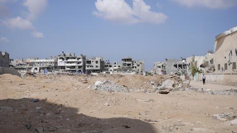 Im Hintergrunde eines öden und leeren Platzes sieht man die Ruinen zerstörter Häuser.