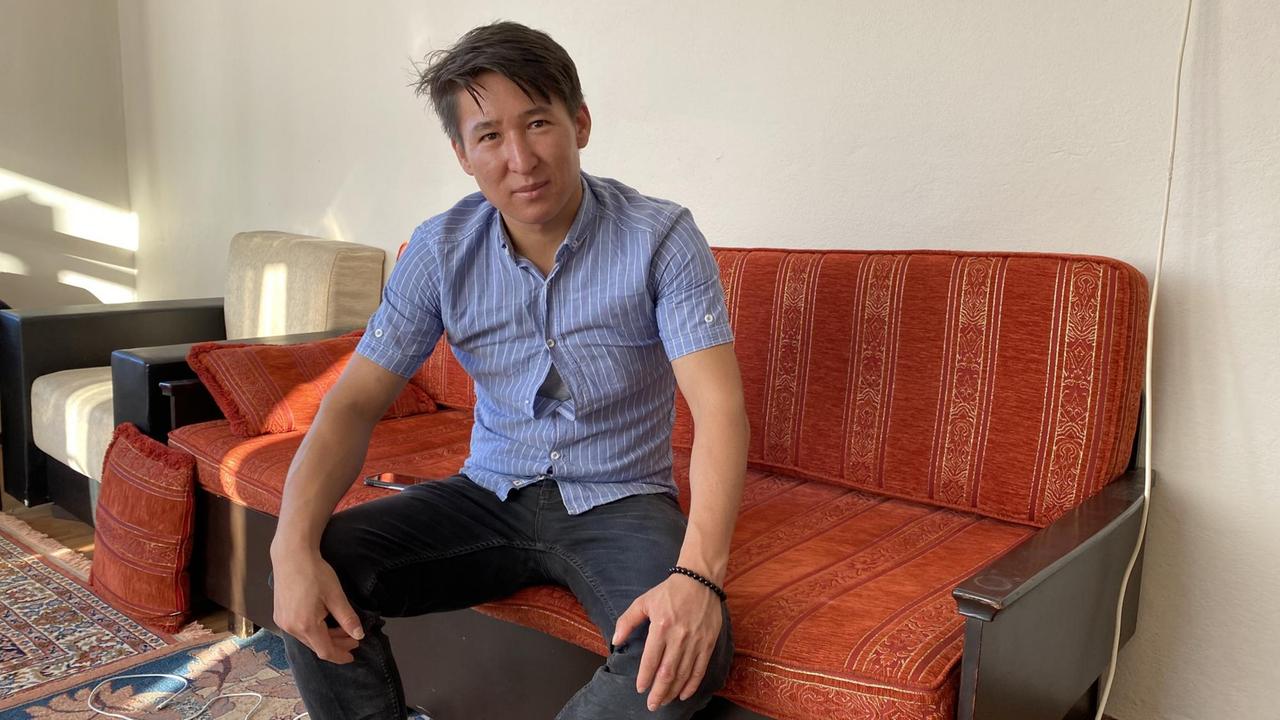 Ein junger schwarzhaariger Mann sitzt breitbeinig in Jeans auf einem roten Sofa und stützt die Hände auf die Knie. Ein junger schwarzhaariger Mann sitzt breitbeinig in Jeans auf einem roten Sofa und stützt die Hände auf die Knie.