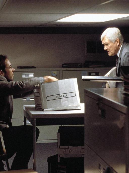 In einem dunkel und äußerst beengt wirkendesn Büro mit tief liegender Decke sprechen John Cusack und Orson Bean über einen Tisch hinweg miteinander.