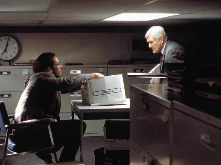 In einem dunkel und äußerst beengt wirkendesn Büro mit tief liegender Decke sprechen John Cusack und Orson Bean über einen Tisch hinweg miteinander.