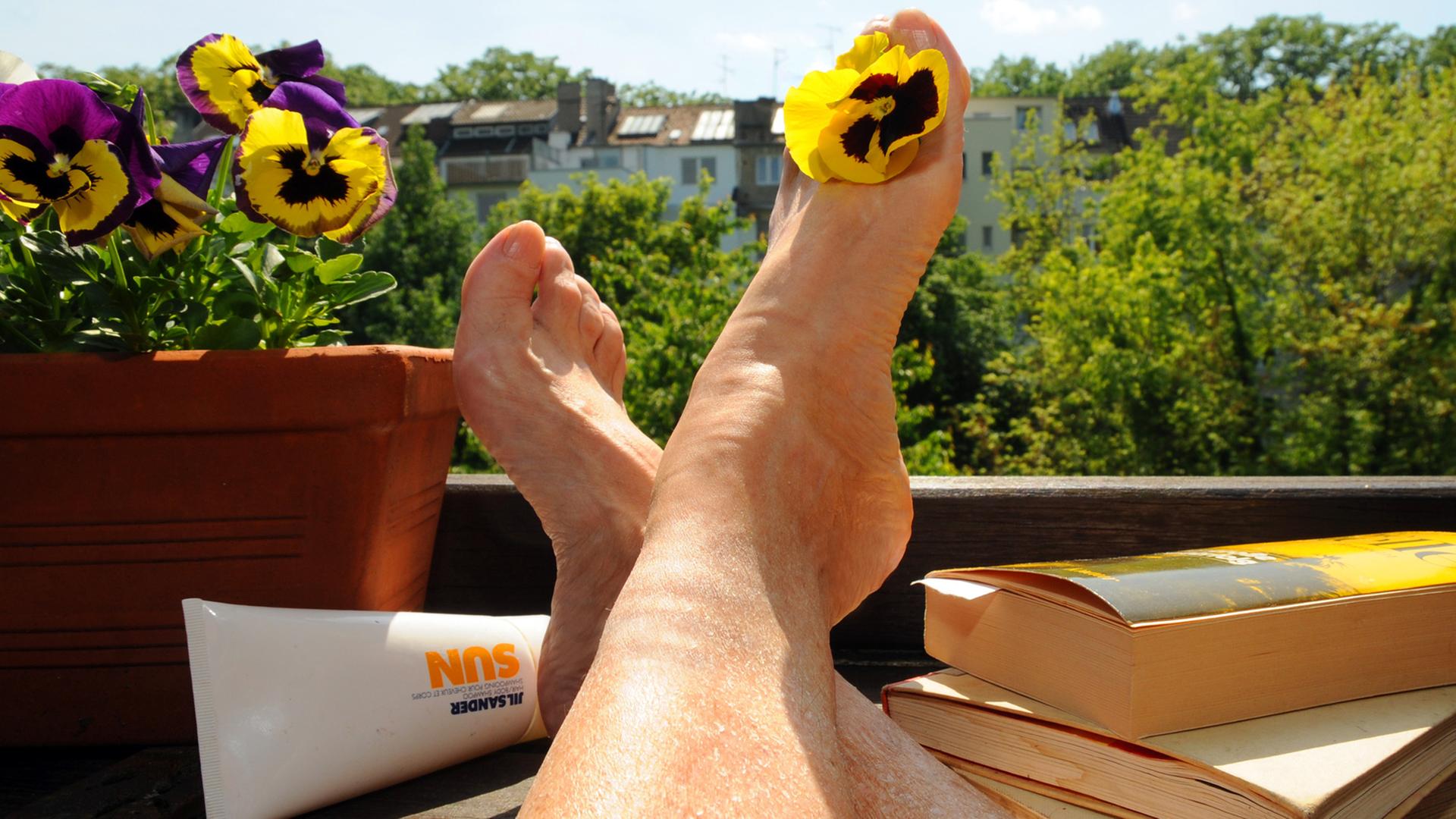 Eine Frau sitzt mit Büchern und Sonnencreme auf einem Balkon und streckt ihre Beine der Sonne entgegen. Eine Frau sitzt mit Büchern und Sonnencreme auf einem Balkon und streckt ihre Beine der Sonne entgegen.