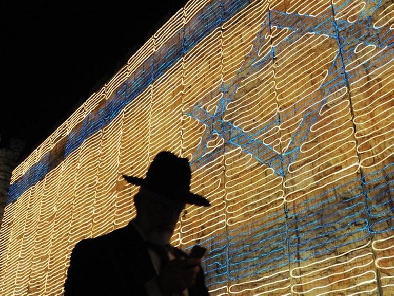 Ein Mann mit Hut vor der israelischen Flagge, bestehend aus Lichterketten an der Wand der alten Stadt Jerusalem