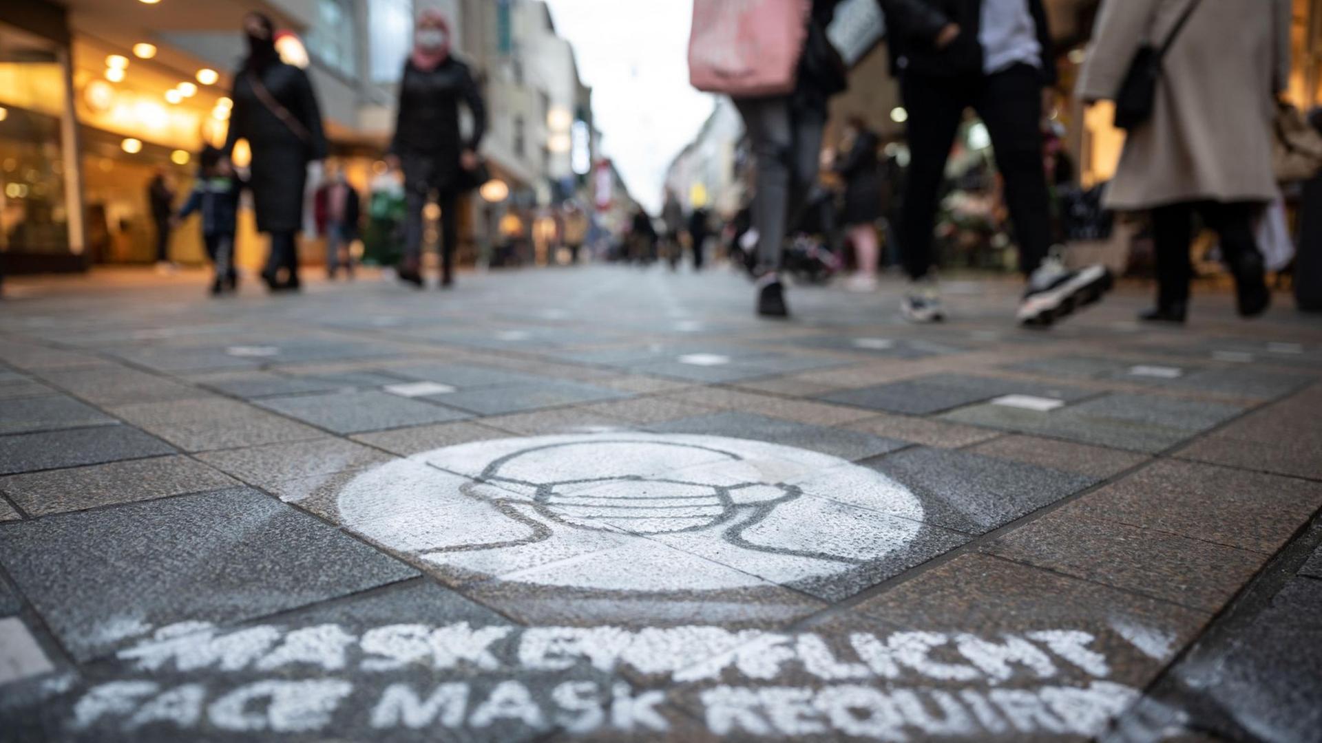 Ein auf den Boden gespraytes Symbol weist in der Fußgängerzone auf die Pflicht hin, Mund-Nasenschutz zu tragen. Ein auf den Boden gespraytes Symbol weist in der Fußgängerzone auf die Pflicht hin, Mund-Nasenschutz zu tragen.