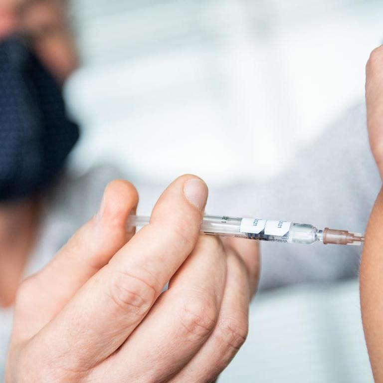 Im Hintergrund sieht man verschwommen einen Mann mit Mundschutz, im Vordergrund groß seine Hand mit einer Spritze und einen kleinen Teil des Oberarms der Patientin