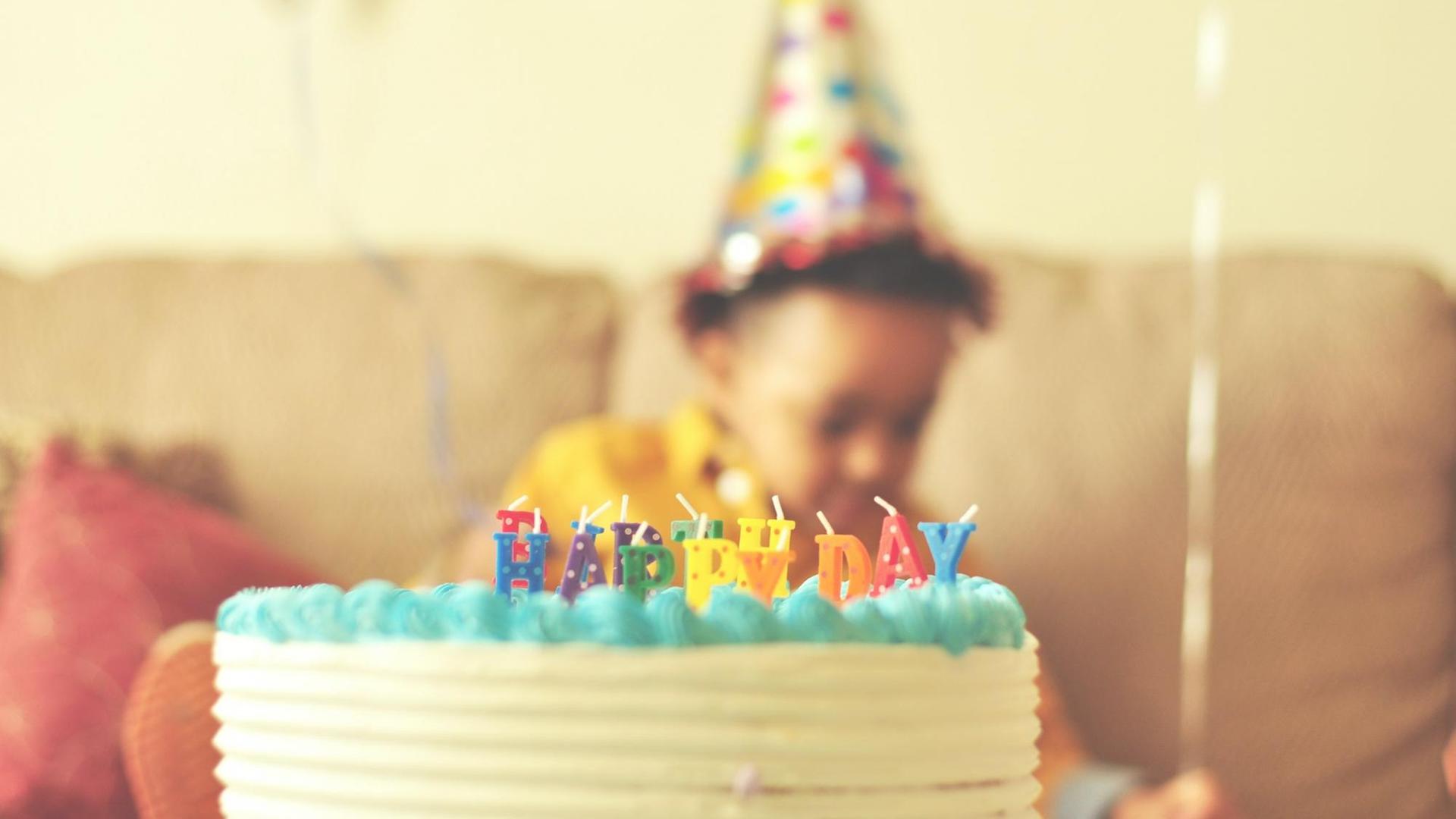 Eine Geburtstagstorte mit "Happy Birthday" in Kerzenbuchstaben. Im Hintergrund ein Kind mit Partyhut. Eine Geburtstagstorte mit "Happy Birthday" in Kerzenbuchstaben. Im Hintergrund ein Kind mit Partyhut.