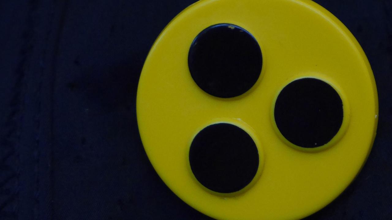 Ein Blindenbutton: Drei schwarze Punkte auf gelbem Kreis. 