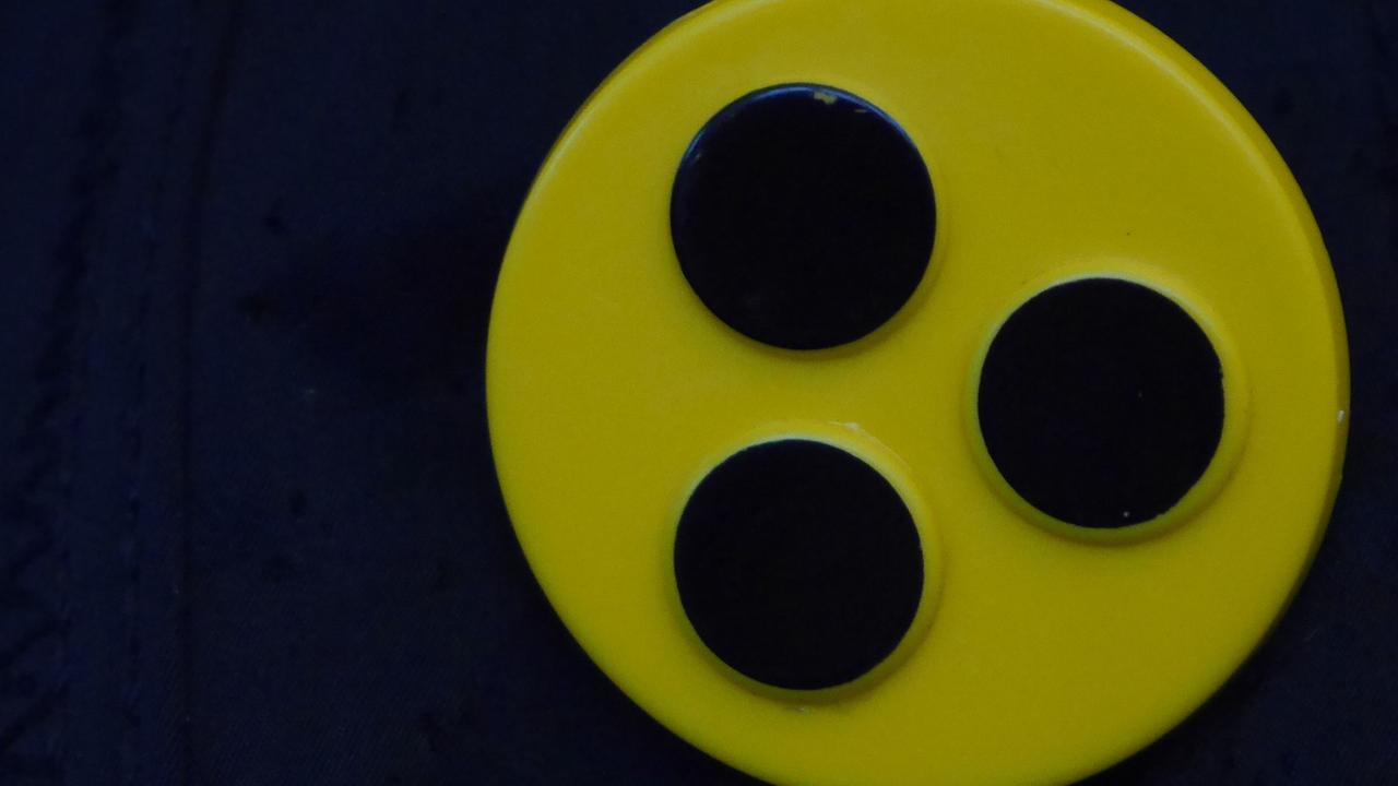 Ein Blindenbutton: Drei schwarze Punkte auf gelbem Kreis. Ein Blindenbutton: Drei schwarze Punkte auf gelbem Kreis.