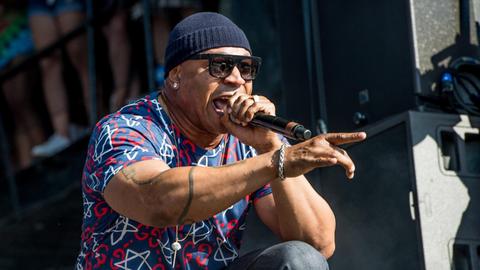 LL Cool J kniet auf der Bühne, rappt und zeigt mit seinem rechten Zeigefinger auf das Publikum. Er trägt Mütze, Sonnenbrille und ein bunt gemustertes Hemd.
