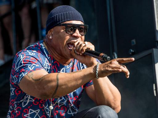 LL Cool J kniet auf der Bühne, rappt und zeigt mit seinem rechten Zeigefinger auf das Publikum. Er trägt Mütze, Sonnenbrille und ein bunt gemustertes Hemd.