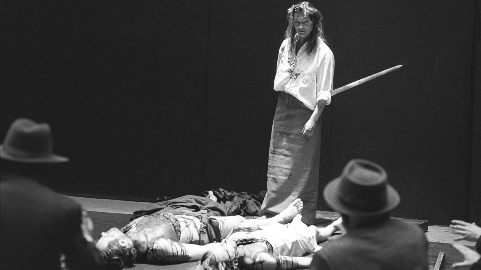 Edith Clever Klytämnestra in einer Inszenierung von Peter Stein an der Schaubühne Berlin 1980. Vor ihr liegen die Leichen von Kassandra und Agamemnon. Im Vordergrund sind umrissartig zwei Männer mit Hüten zu erkennen.