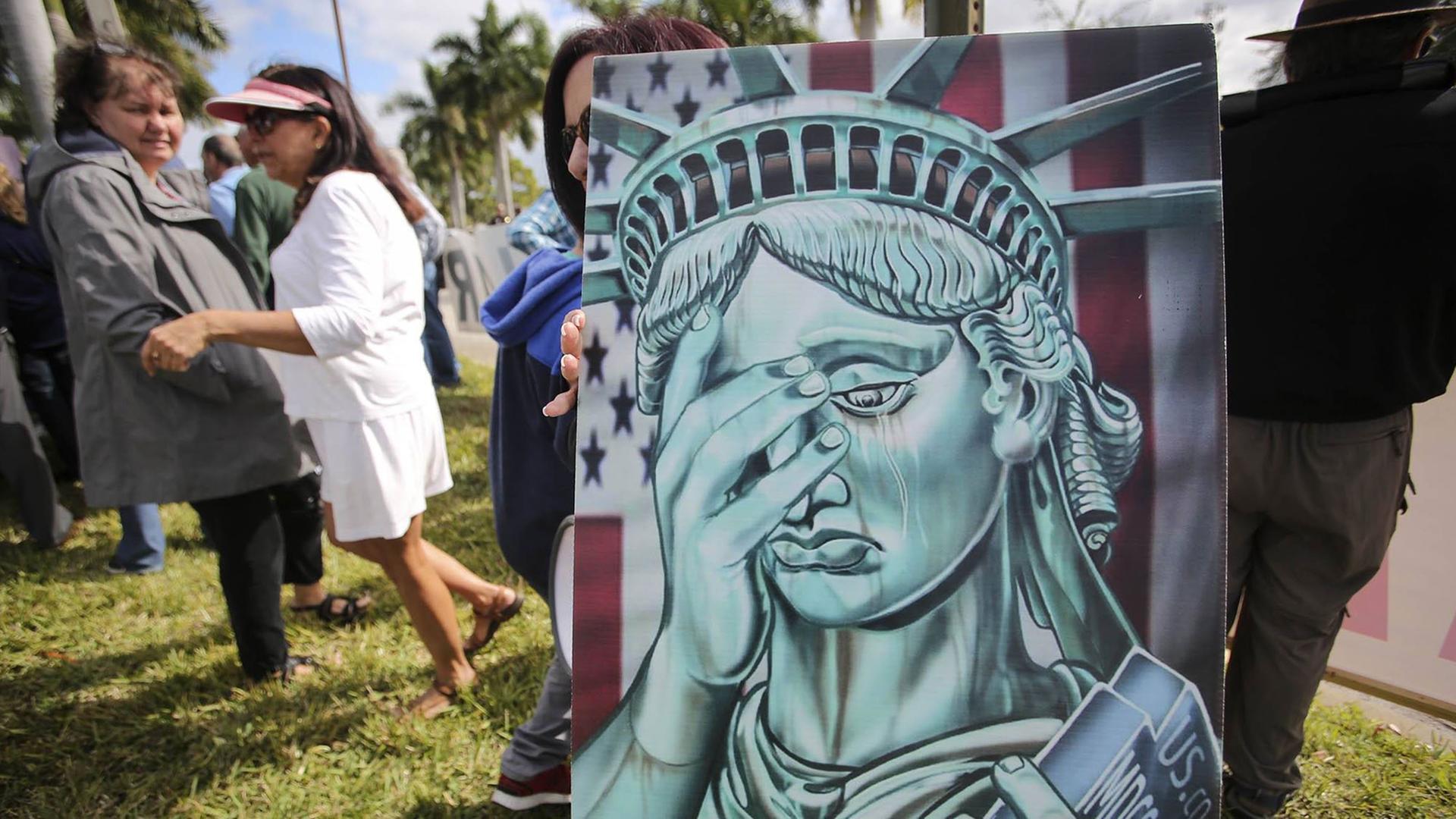 Zu sehen sind Menschen am 21. Januar 2019 vor dem Gemeindehaus in West Palm Beach, Florida. Jemand hält dabei ein Bild mit der Statue of Liberty, die sich eine Hand vor das Gesicht hält. Zu sehen sind Menschen am 21. Januar 2019 vor dem Gemeindehaus in West Palm Beach, Florida. Jemand hält dabei ein Bild mit der Statue of Liberty, die sich eine Hand vor das Gesicht hält.