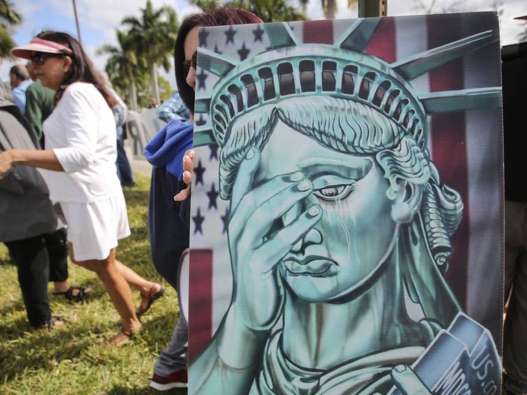 Zu sehen sind Menschen am 21. Januar 2019 vor dem Gemeindehaus in West Palm Beach, Florida. Jemand hält dabei ein Bild mit der Statue of Liberty, die sich eine Hand vor das Gesicht hält.