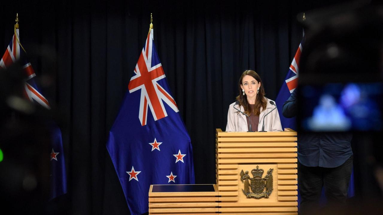 Jacinda Ardern steht an einem Pult und spricht, im Hintergrund ist die Neuseeländische Flagge zu sehen. Jacinda Ardern steht an einem Pult und spricht, im Hintergrund ist die Neuseeländische Flagge zu sehen.