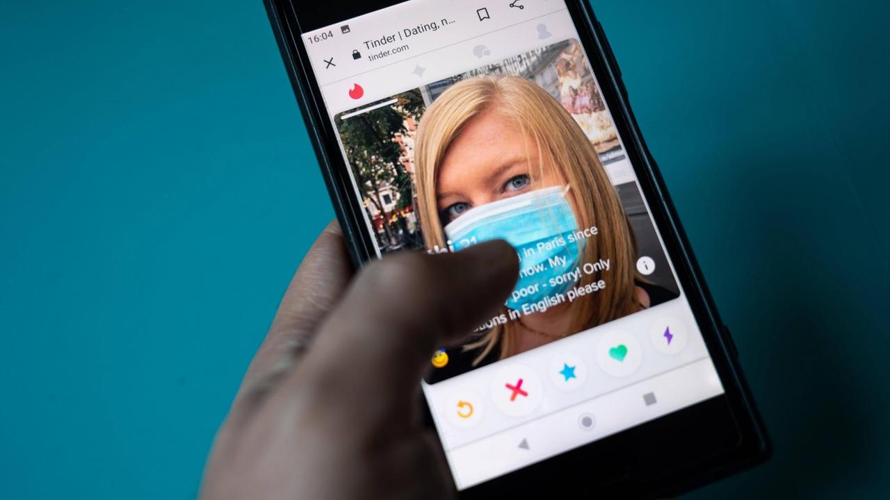 Eine Hand hält ein Smartphone mit der Tinderapp; angezeigt wird dort gerade eine blonde Frau mit Corona-Schutzmaske Eine Hand hält ein Smartphone mit der Tinderapp; angezeigt wird dort gerade eine blonde Frau mit Corona-Schutzmaske
