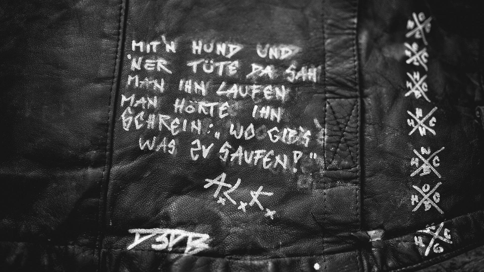 Auf einer dunklen Lederjacke ist mit weißem Edding der Spruch "Mit'n Hund und 'ner Tüte, da sah man ihn laufen. Man hörte ihn schrein: Wo gib's was zu saufen? ALK" aufgemalt. Auf einer dunklen Lederjacke ist mit weißem Edding der Spruch "Mit'n Hund und 'ner Tüte, da sah man ihn laufen. Man hörte ihn schrein: Wo gib's was zu saufen? ALK" aufgemalt.