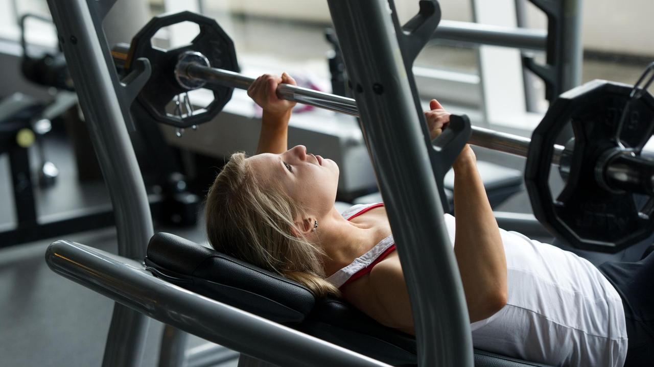 Eine junge Frau in einem Fitnessstudio drückt eine Langhantel auf der Schrägbank in die Höhe. Eine junge Frau in einem Fitnessstudio drückt eine Langhantel auf der Schrägbank in die Höhe.