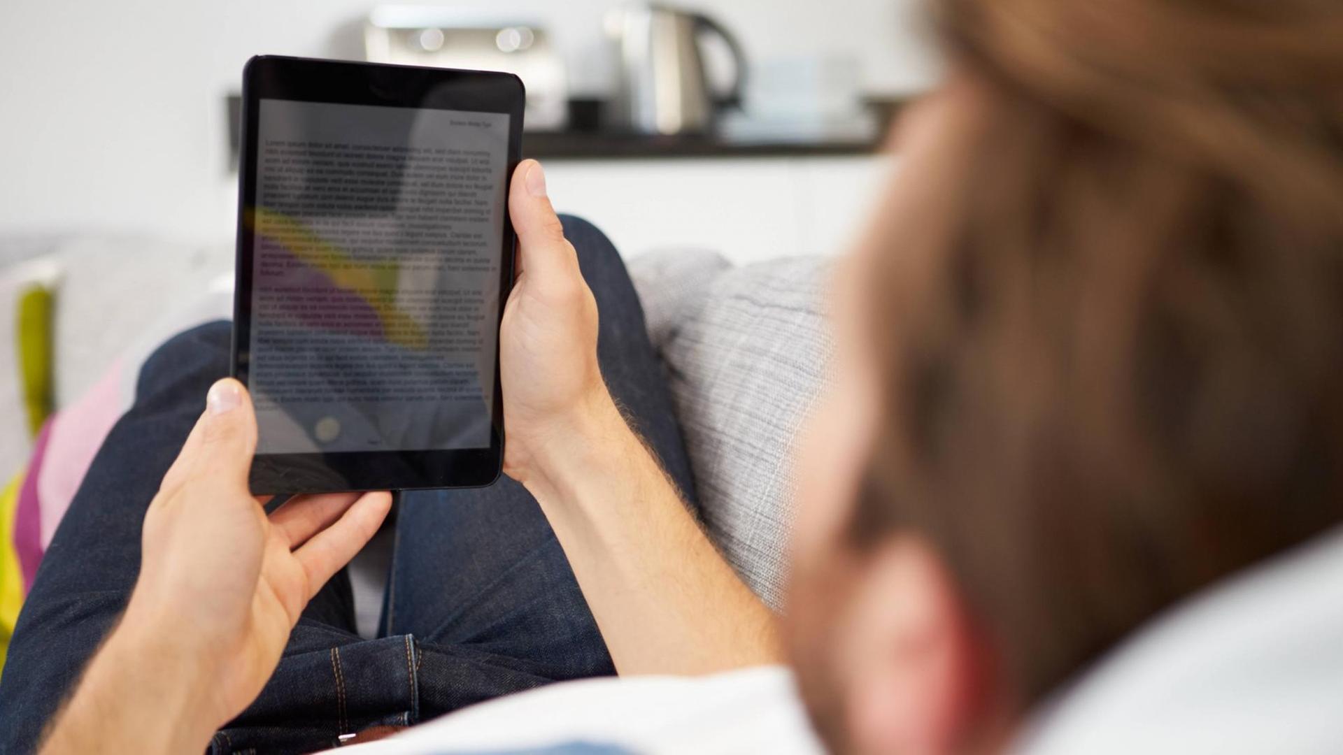 Ein junger Mann liegt auf dem Sofa und hält einen Ebook-Reader in den Händen. Ein junger Mann liegt auf dem Sofa und hält einen Ebook-Reader in den Händen.