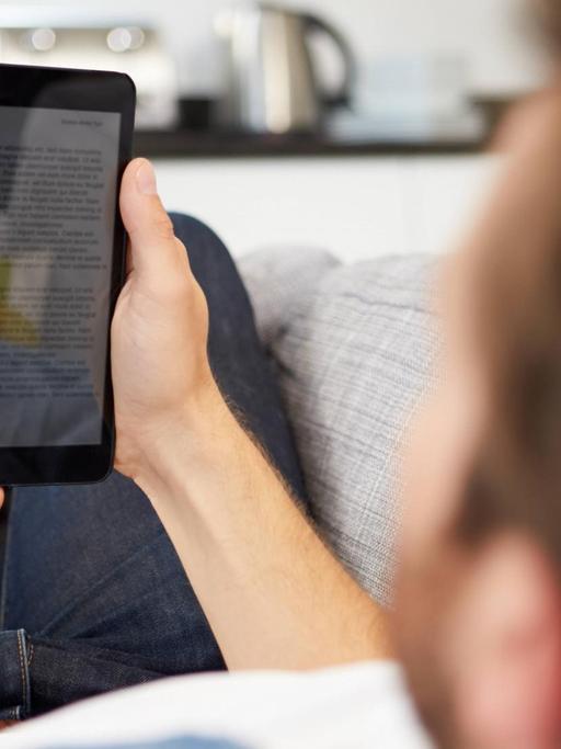 Ein junger Mann liegt auf dem Sofa und hält einen Ebook-Reader in den Händen.