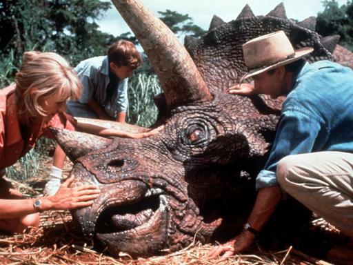 Die Schauspieler Laura Dern, Joseph Mazzelli und Sam Neil helfen einem kranken Dinosaurier. (Aufnahme von 1993). Im Film "Jurassic Park" experimentieren Wissenschaftler mit dem Erbgut von Dinosauriern, das sie in konservierten, blutsaugenden Insekten aus der Vorzeit entdecken. Es gelingt ihnen, selbst Dinosaurier heranzuzüchten, die später ihre Schöpfer terrorisieren.