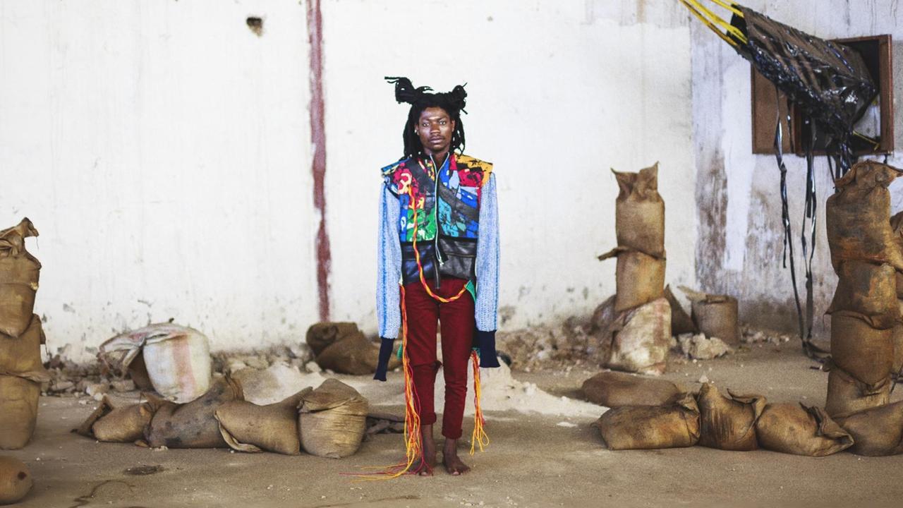 Ein männliches Model präsentiert in einer etwas heruntergekommenen Lagerhalle zwischen Säcken unbekannten Inhalts eine bunte Jacke mit überlangen Ärmeln von der Designerin Njola Impressions. Ein männliches Model präsentiert in einer etwas heruntergekommenen Lagerhalle zwischen Säcken unbekannten Inhalts eine bunte Jacke mit überlangen Ärmeln von der Designerin Njola Impressions.