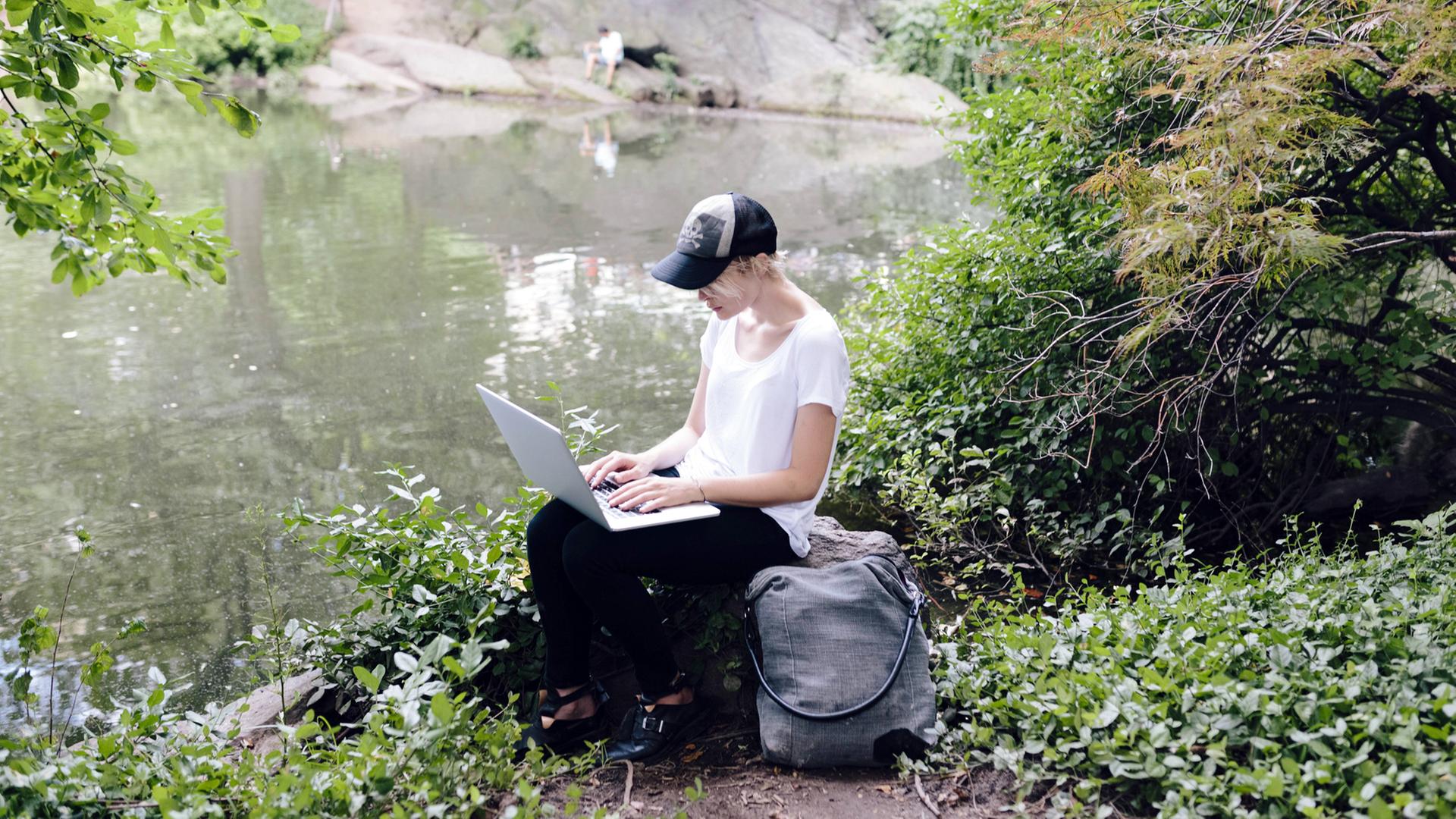 Eine junge Frau sitzt mit ihrem Laptop an einem Seeufer im Grünen Eine junge Frau sitzt mit ihrem Laptop an einem Seeufer im Grünen